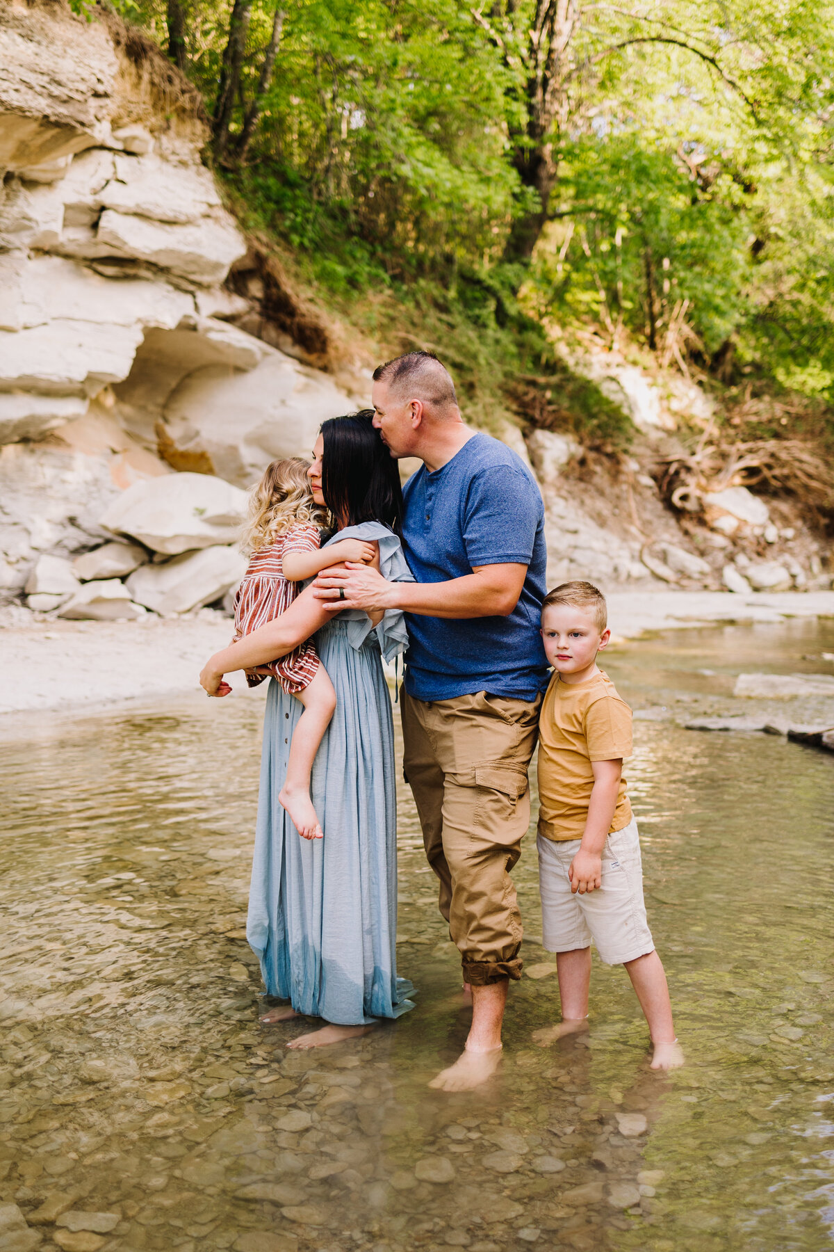 Family photo in a river with rocks and trees around them, the woman is carrying the girl with a flower dress and the man is kissing her on the head with brown pants and blue shirt and the boy is hugging his father's leg with a yellow t-shirt