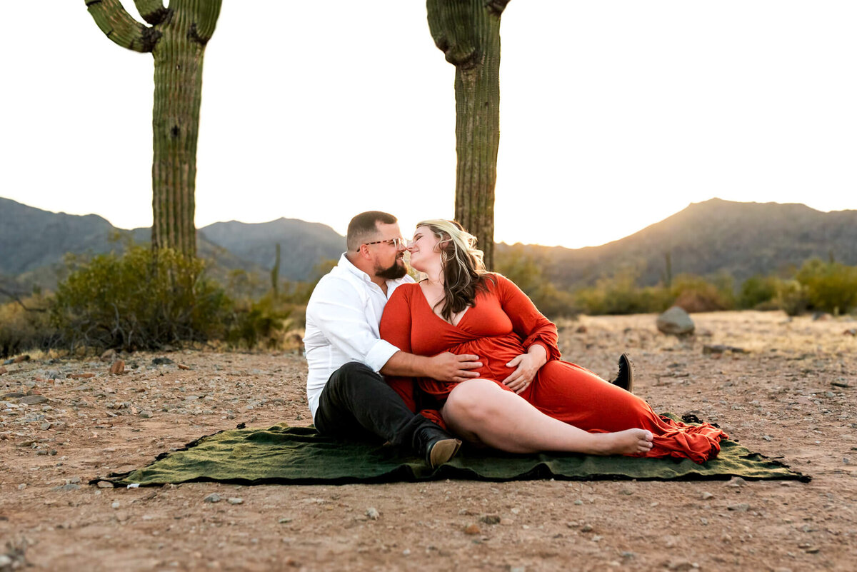 Expecting parents sit on a picnic blanket amid the Arizona desert and lean in for a kiss