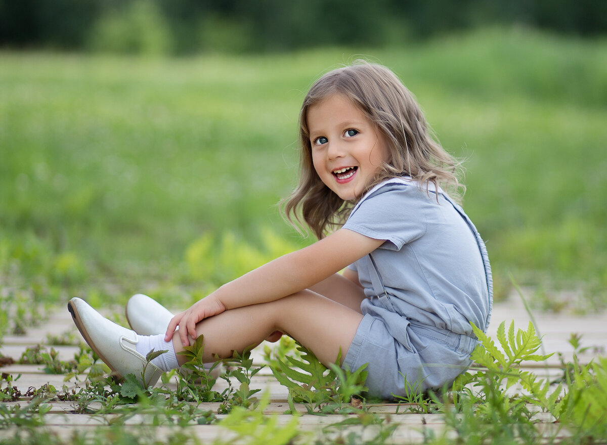 Little boy is sitting in a brooklyn park at a photoshoot  to commemorate his upsherin. He is looking over his shoulder and smiling at the camera. Captured by Chaya Bornstein, Brooklyn Family Photographer