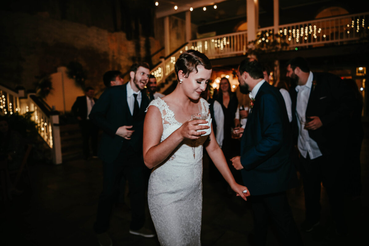 A bride in a white lace gown holds a drink and smiles while dancing at a dimly lit wedding reception with guests in formal attire around her. Twinkling string lights decorate the background.