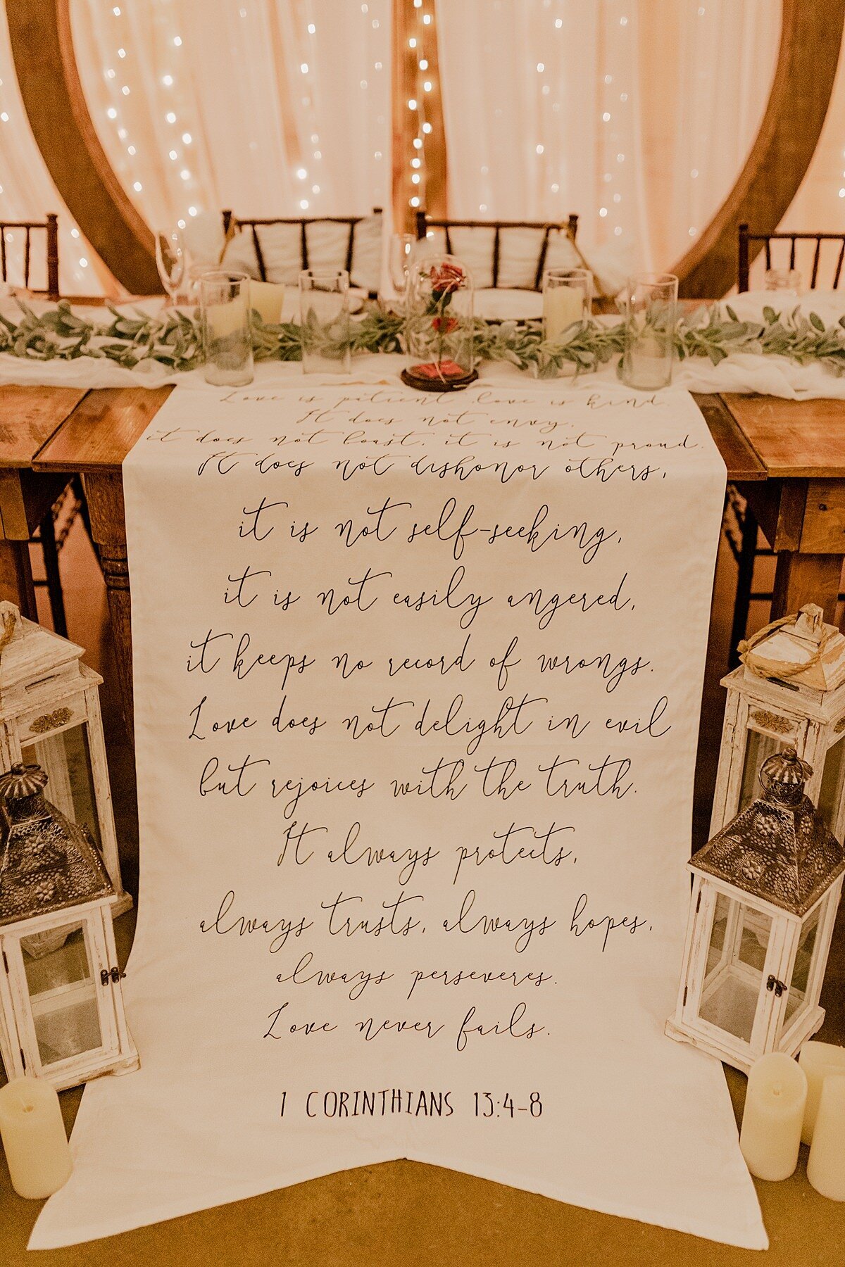 A banner with 1 Corinthians 13.4-8 decorates a sweetheart table at Grace Valley Farm. On either side of the banner are two sets of white lanterns with pillar candles. The farm house tables are decorated with white organza table runners with a lambs ear garland and clear glass cylinders with white pillar candles. Behind the table is a dark wood round arbor with sheer drapery and twinkle lights.