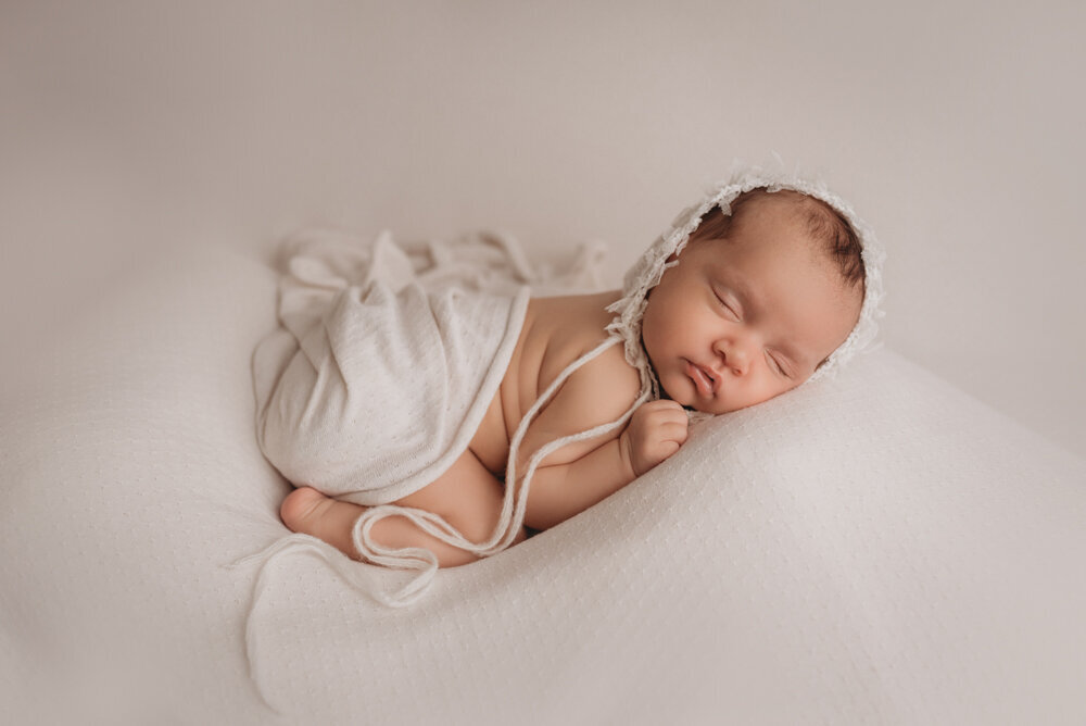 Newborn photo shoot photographed by Atlanta, Georgia newborn photographer with baby posing on neutral fabric with dainty headband on and sleeping. Baby is laying on her tummy wearing a white hat.
