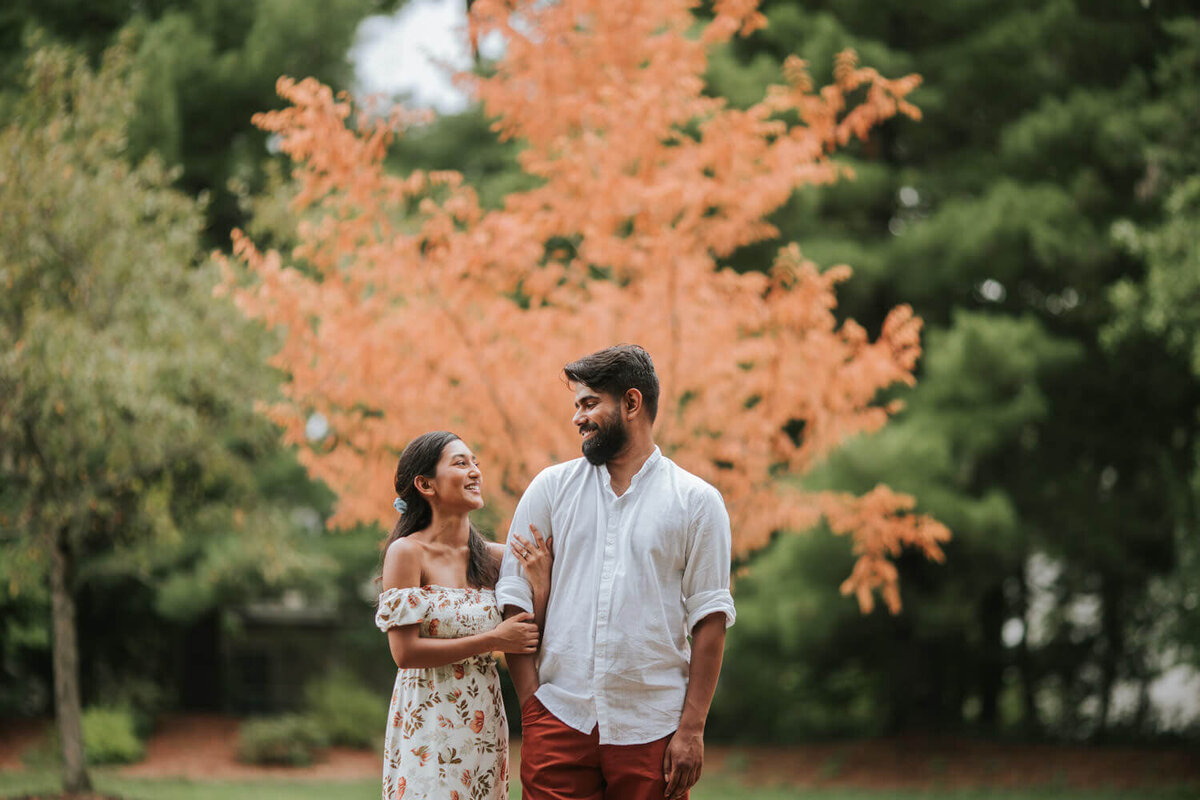 A couple shares a joyful moment in Hamilton Square, NJ, with a vibrant orange tree creating a striking autumnal backdrop.