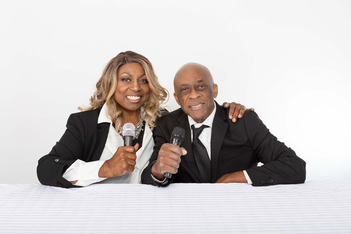 A charismatic couple in coordinating black suits pose together at a white table holding microphones, smiling warmly for a personal branding session tailored for podcast hosts or speakers.