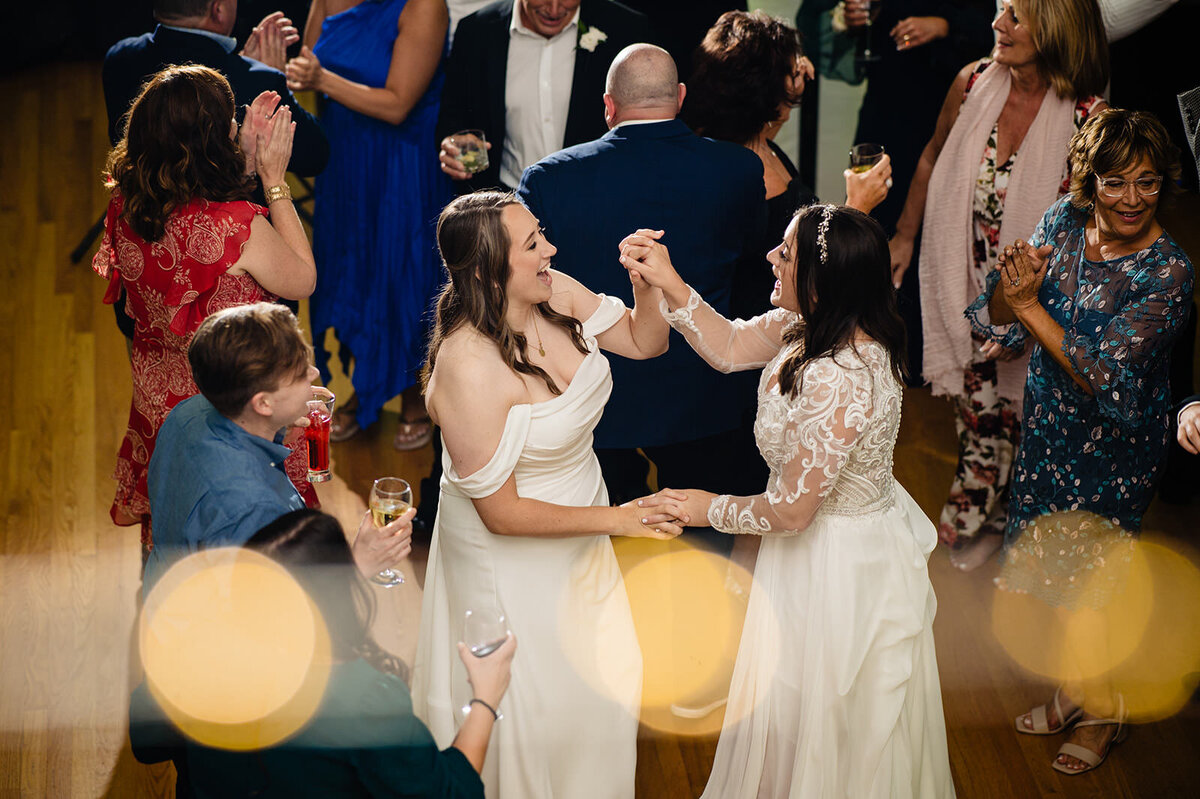 A bird's-eye view of the brides on the dance floor, with one bride seated and the other reaching out to her, surrounded by dancing guests and soft lighting.