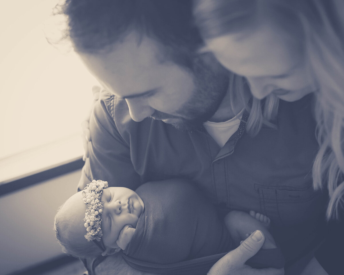 New parents smile down at their sleeping baby daughter in dad's arms under a window