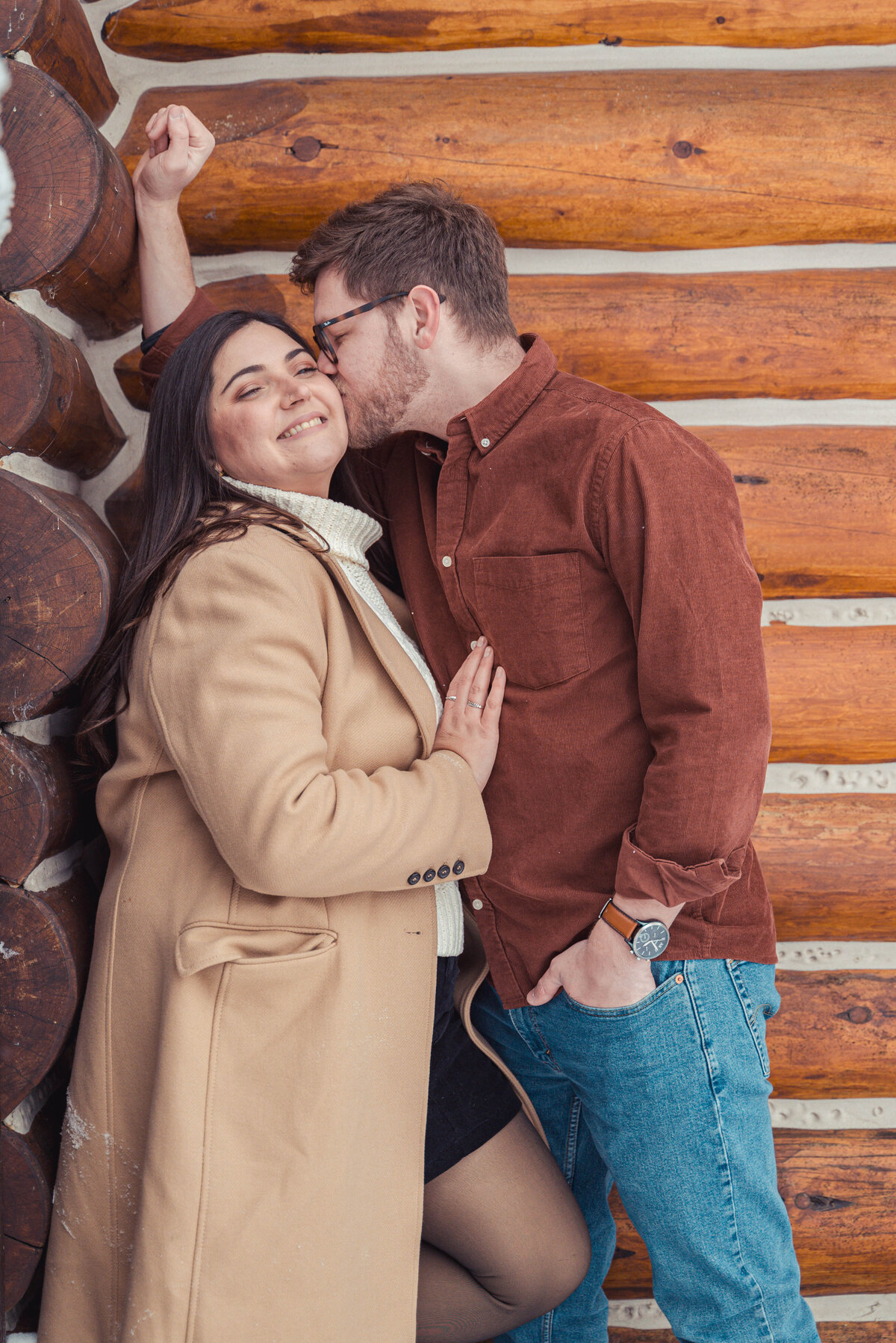 Couple cuddles against rustic log cabin during cozy Banff winter session.
