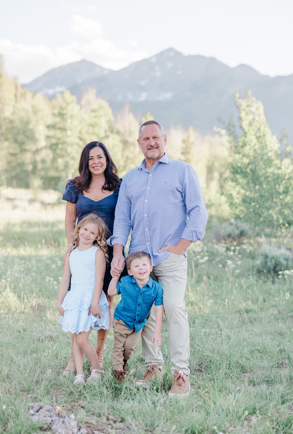 A grandma and grandpa smile while standing with their toddler grandkids at their feet