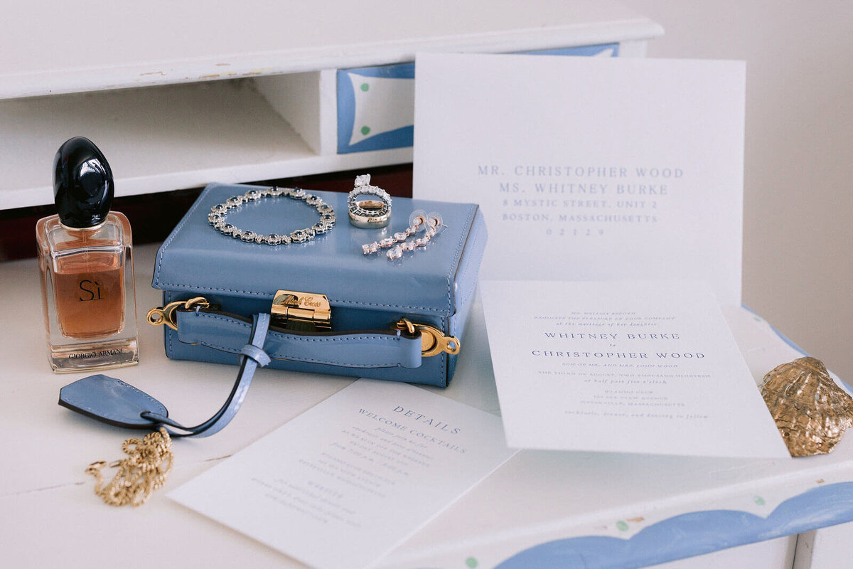 Powder blue purse with a bracelet, rings, and earrings on top of it, a perfume, and a wedding invitation at Cape Cod, MA.