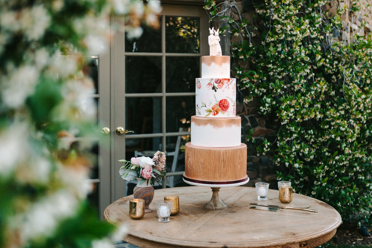 A photograph in color of a wedding cake, during the reception at Saddlerock Ranch in Malibu, California. The wedding cake is on a round wooden table surrounded by blooming jasmine and french doors of a building in the background. The cake is four tiers with a white ceramic cake topper of two rabbits standing arm in arm. The bottom tier of the cake is painted like a stump of wood, which matches the tops of the first and third tiers. The second tier from the top is painted with orange and pink florals. Wedding photography by Stacie McChesney/Vitae Weddings.