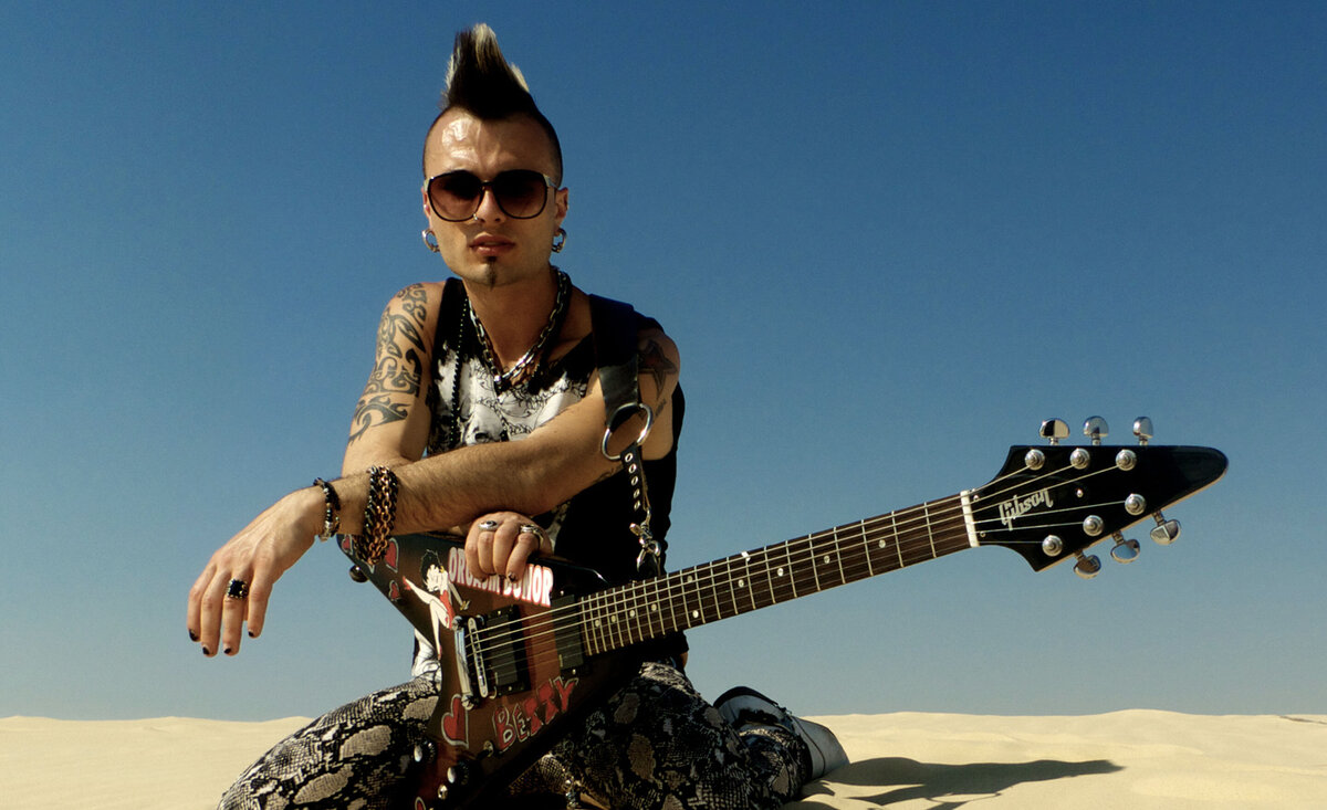 Male musician photo Sal Costa wearing black and white outfit kneeling with electric guitar against sand dune background