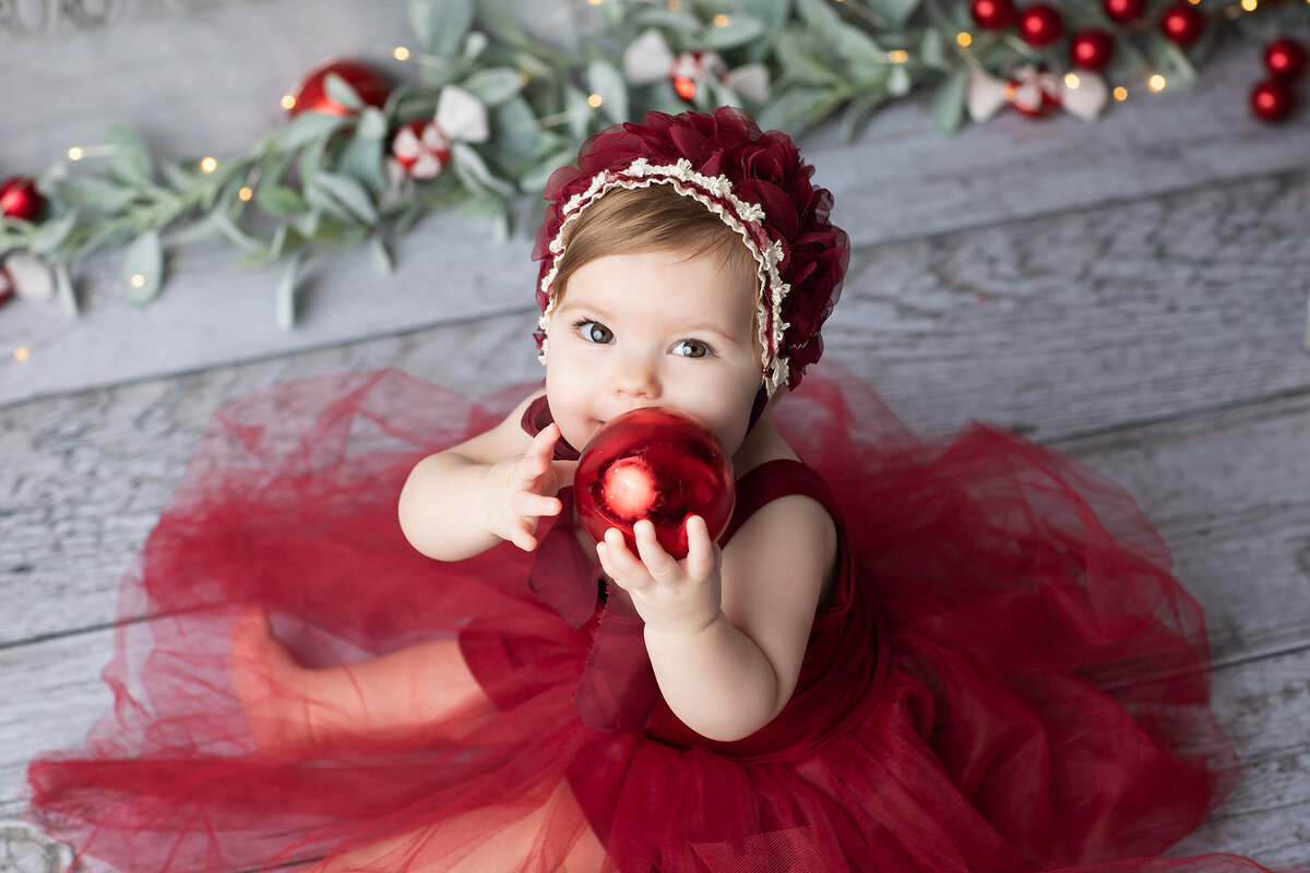Baby girl in a burgundy holiday dress and matching bonnet holding a shiny red ornament, surrounded by festive greenery and lights in a Dallas milestone photographer setup.