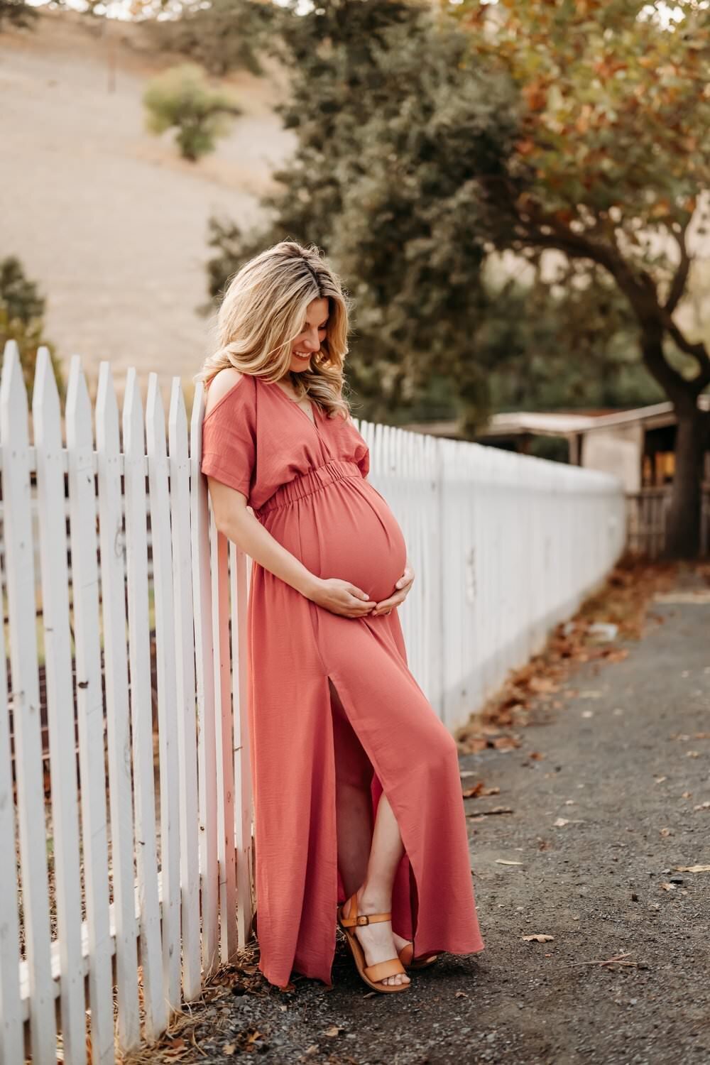 woman in peach maternity dress leans against a white fence.