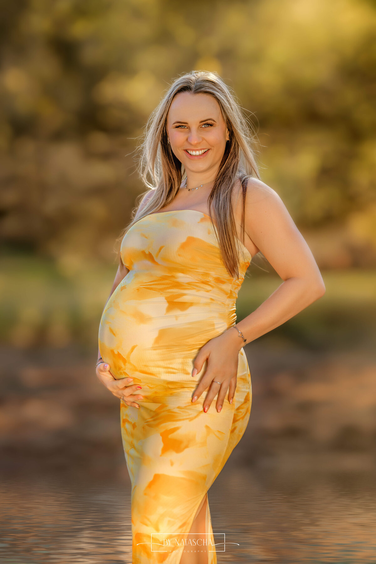 Zwangere vrouw in gele jurk in het bos voor zwangerschapsshoot in Berghem, Herperduin lachend naar de camera