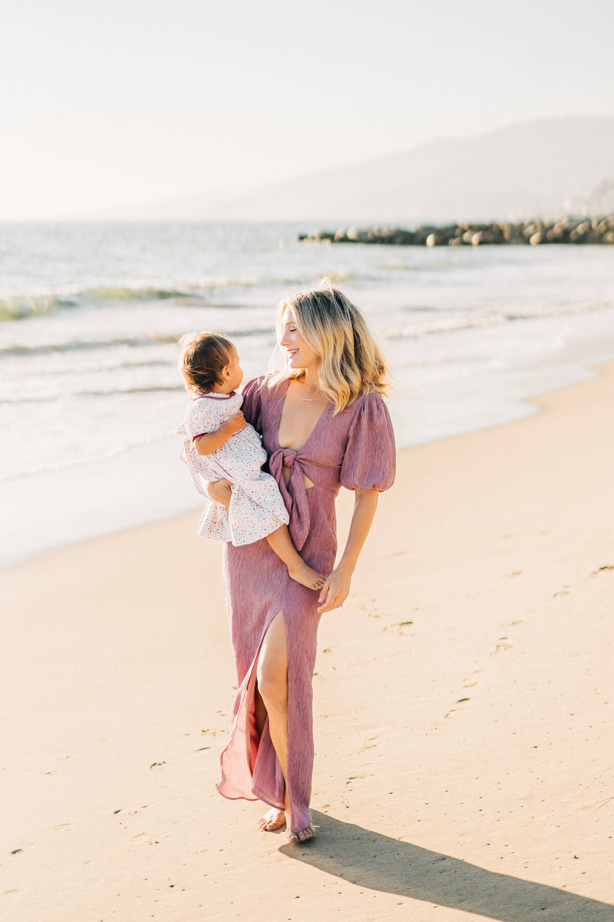 Mom is wearing a purple dress at the beach and holding daughter during their family photography session