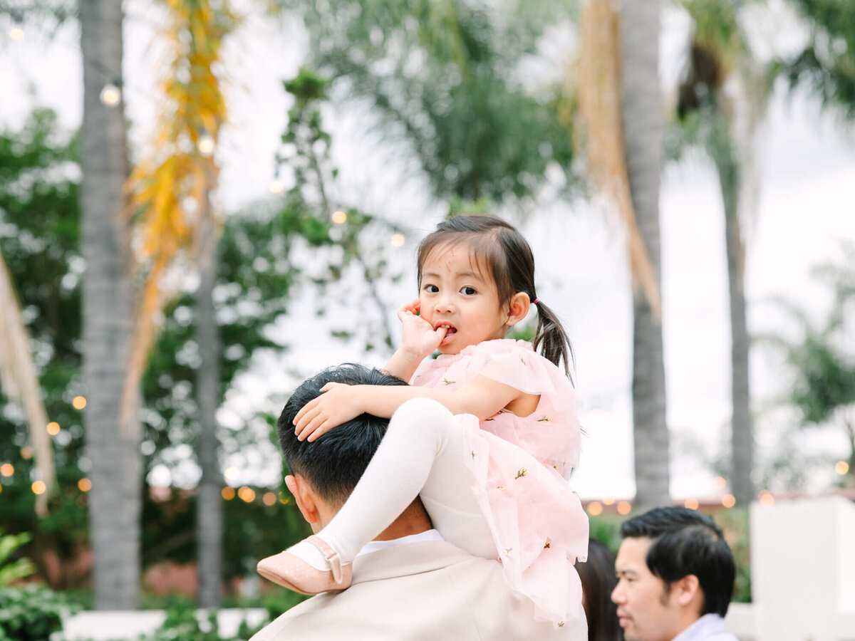 A child in a pink dress sits on a man's shoulders, surrounded by palm trees and string lights. The scene feels joyful and festive outdoors.