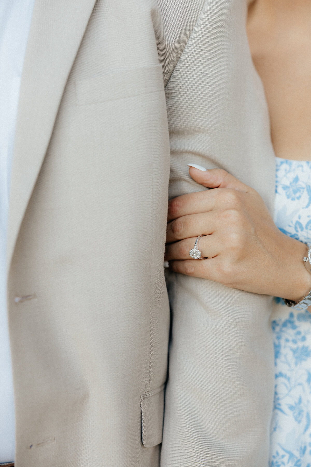 A girl in a light blue floral dress holding the arm of her fiance who wears a light tan jacket.