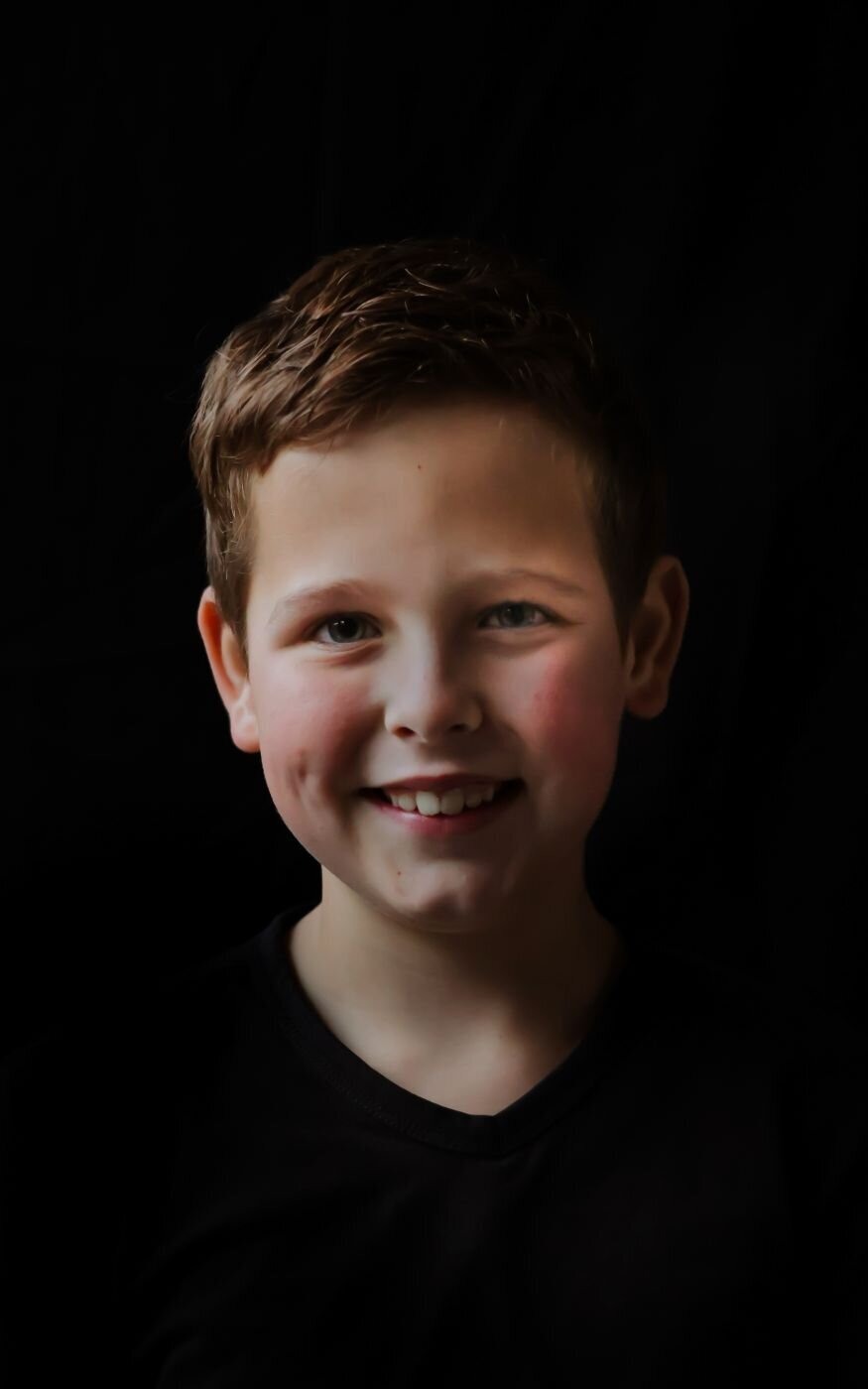 Een portretfoto van een jonge jongen met een open en vriendelijke uitstraling, gefotografeerd tegen een donkere achtergrond