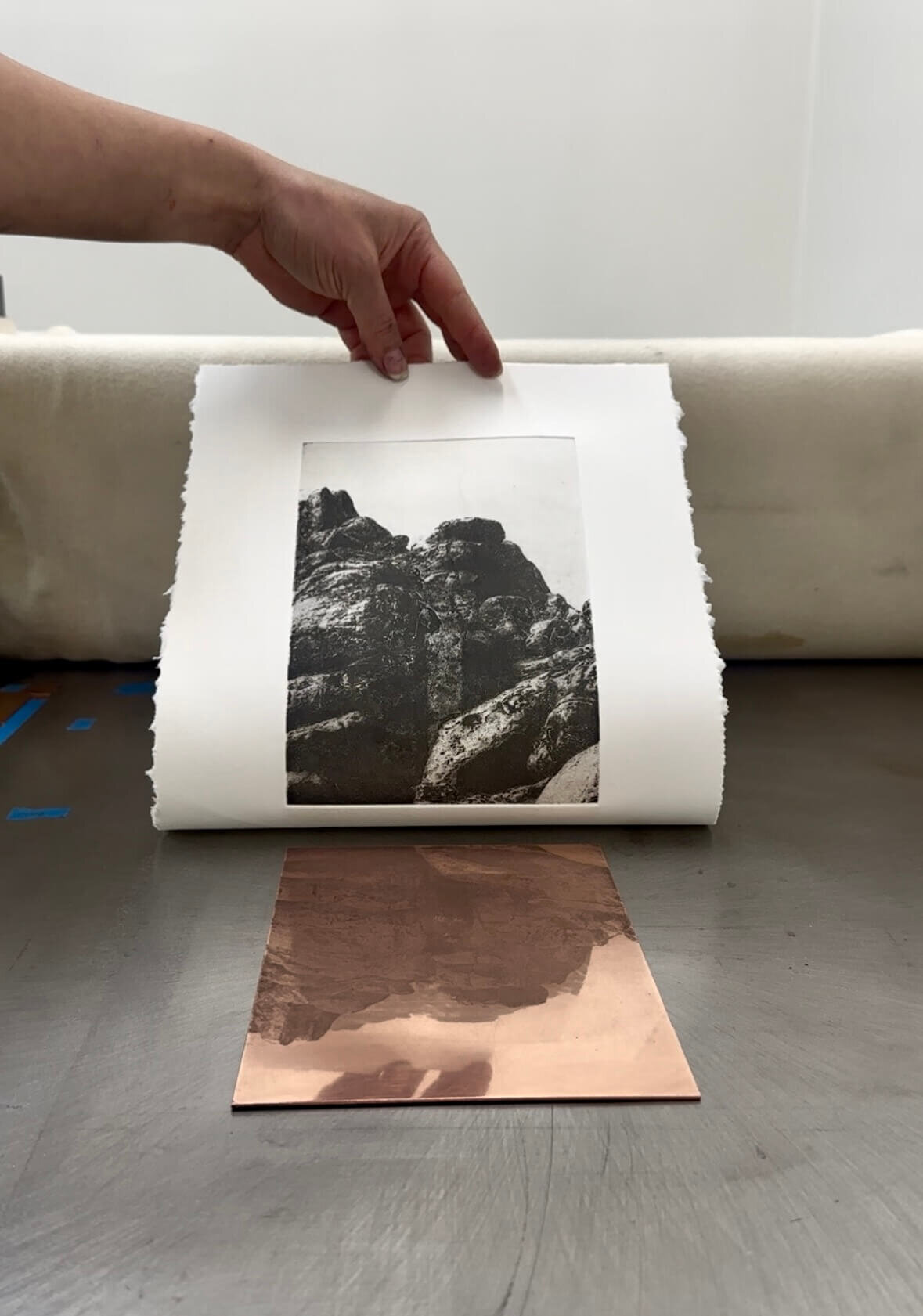 A completed etching print of a rock formation being lifted off a copper plate on the etching press.