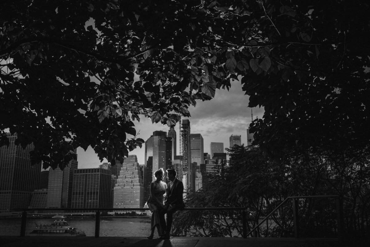 A couple sitting on the railing in a park with a city skyline behind them.