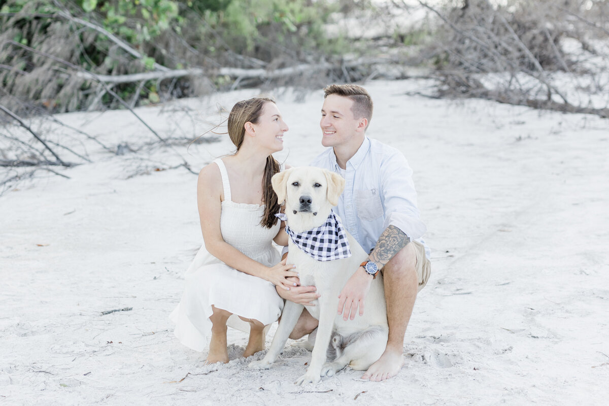 Beach engagement photos in Florida with a yellow lab dog.  Their cute pup is wearing a blue bandana and looking right at the camera while the couple is looking at each other.