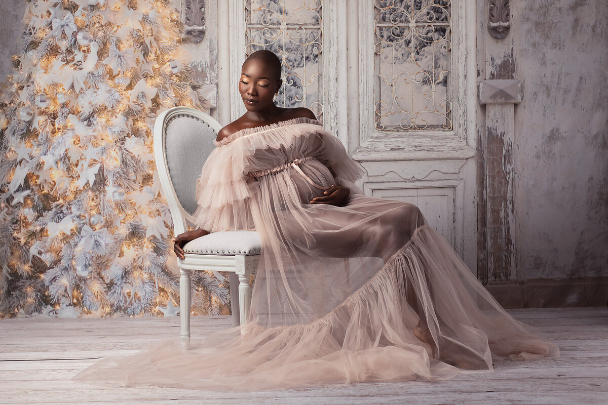 A serene setting featuring an expecting mother dressed in a delicate blush tulle gown, seated gracefully on a vintage chair beside a glowing, frosted Christmas tree. This beautiful photograph is captured by talented Dallas maternity photographer Lindsay Walden.