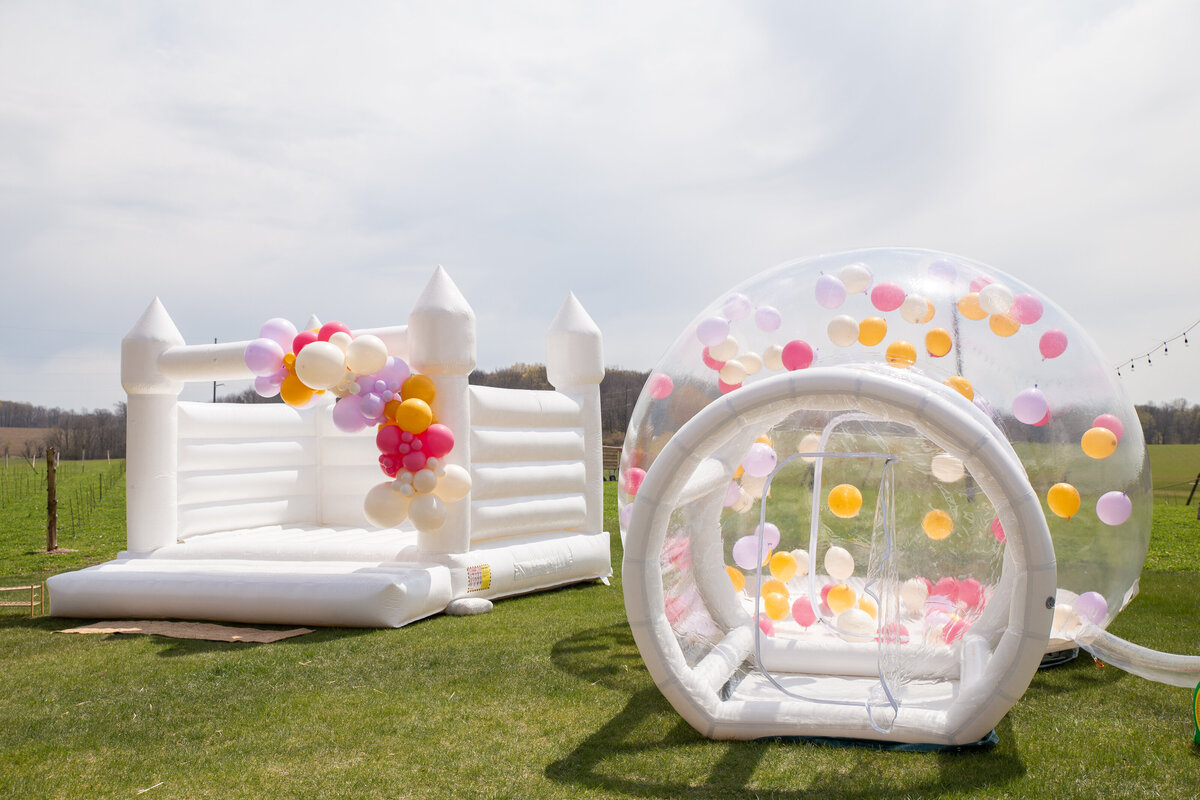 A white bounce house next to a large transparent bubble dome with balloons.