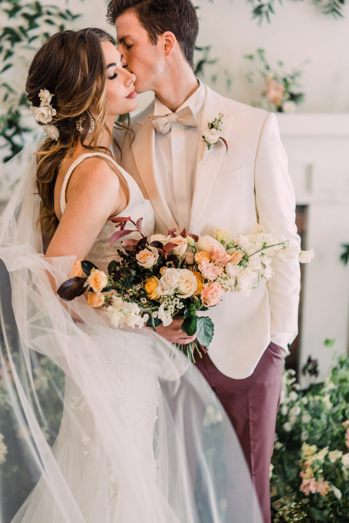 Bride with flowing veil & roses in her hair holding bouquet & closing her eyes while groom in white tuxedo kisses her cheek