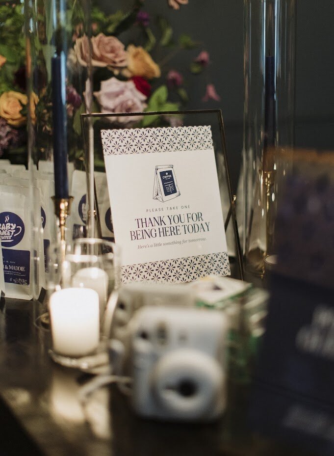 A white thank you sign with navy blue text and line art of a coffee bean bag, expressing gratitude to wedding guests at North & Navy, Ottawa. The thoughtful detail highlights the couple’s coffee bean favors, with navy blue tapered candles, polaroid camera, florals, and tea lights adding charm to the display. This custom wedding signage blends personalization and elegance, reflecting Frid Events' attention to unique, stylish decor and stationery for weddings and special events in Ottawa.