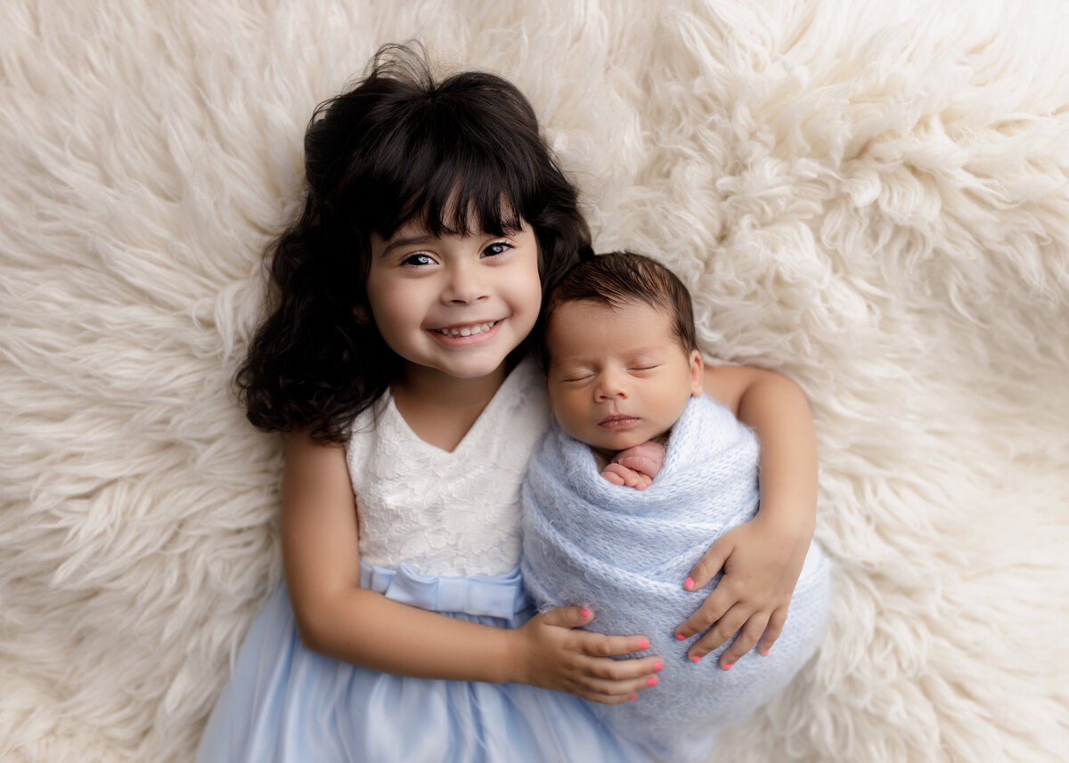 Newborn photoshoot with big sister in West Palm Beach Florida. Big sister is proudly holding her baby brother atop of a fuzzy long faux fur rug. Big sister is smiling at the camera wearing a white and blue dress with baby's head resting atop her shoulder. Baby is in a blue knit wrap with his hands peeking out the top. Aerial image.