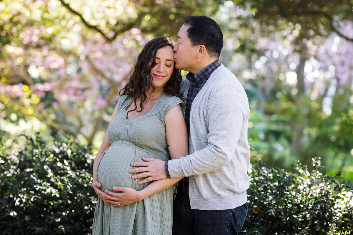 Pregnant woman in light green dress leaning on her husband and closing her eyes as he kisses her at Blake Gardens in Berkeley