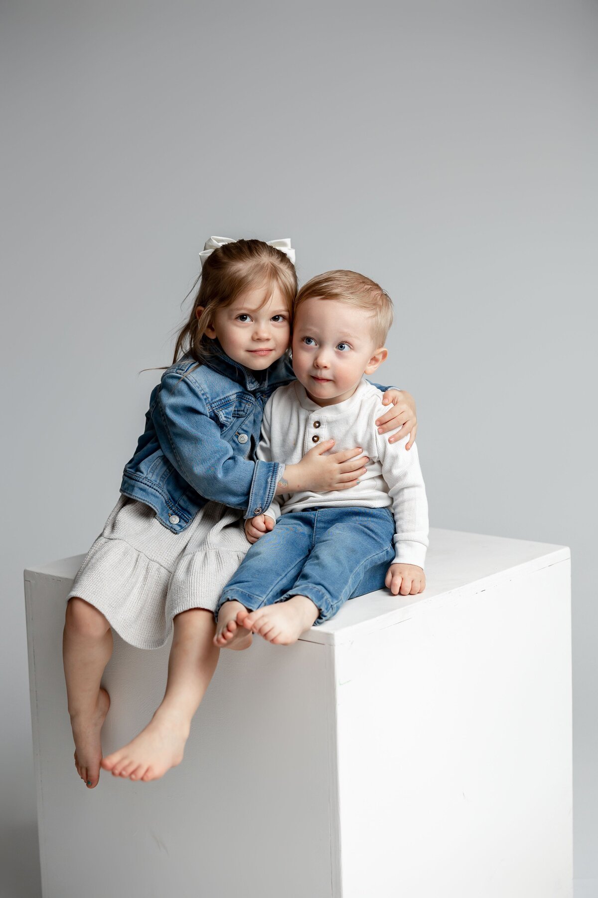Two siblings hugging on white cube, both dressed in matching denim jackets. Photographed by Yucaipa family photographer Kaitlyn Dawn Photography.