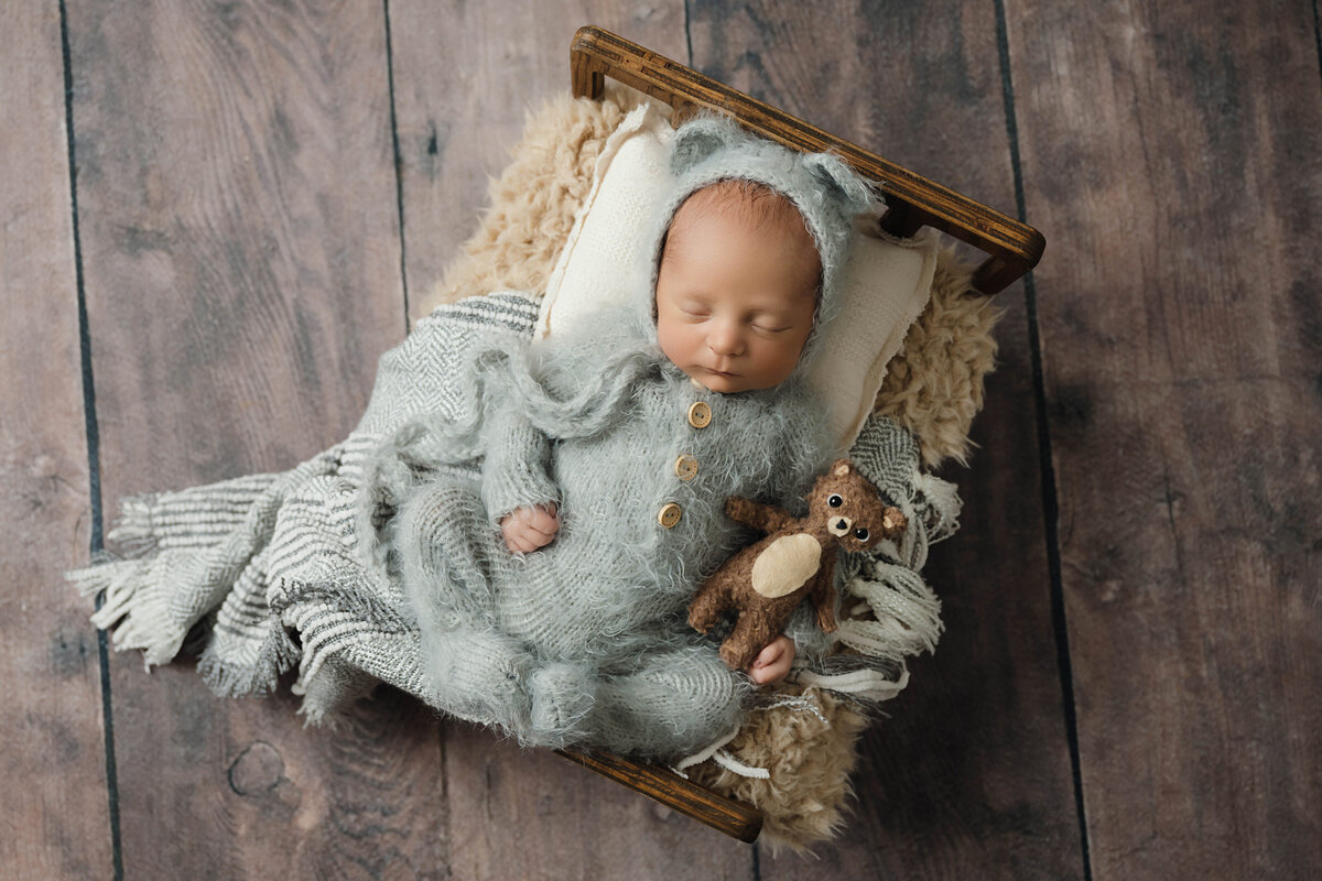 Newborn boy is dressed in a fuzzy grey bear jumper and holding a tiny brown bear while sleeping on a tiny brown bed during his session with the best newborn photographer in Pittsburgh, Katie Louise Photography.