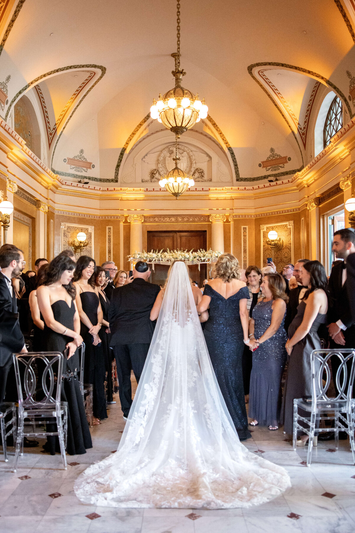 A wedding ceremony in an elegant hall with a high ceiling and ornate chandeliers. A bride in a long white gown and veil stands facing the officiant, surrounded by guests in formal attire. Clear chairs line the aisle.