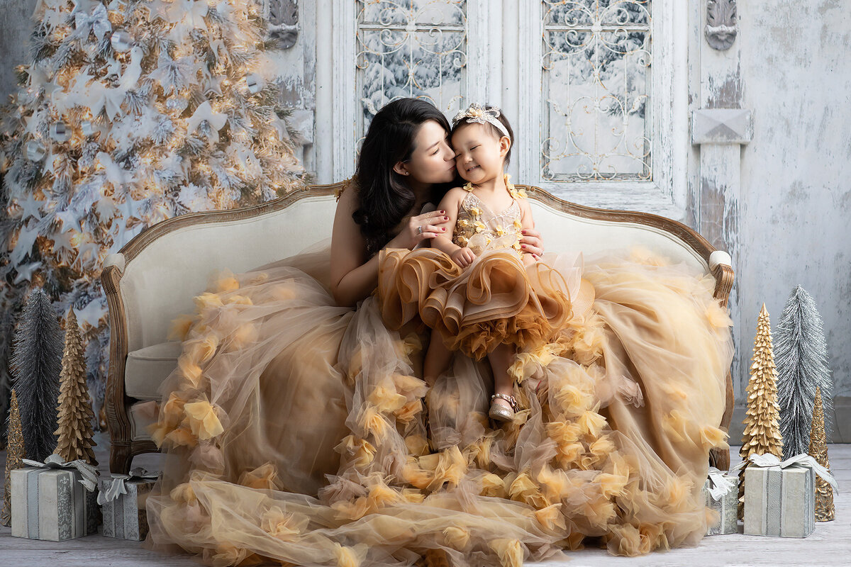 Luxury mother and daughter Christmas portrait in an elegant studio setting. A mother in a flowing gold couture gown kisses her smiling daughter, dressed in a matching ruffled dress, as they sit on a vintage sofa. The backdrop features a frosted holiday tree with gold and silver ornaments, metallic gift boxes, and decorative trees, creating a dreamy winter wonderland. Captured in a fine art photography studio in Dallas, Fort Worth, and surrounding DFW areas.