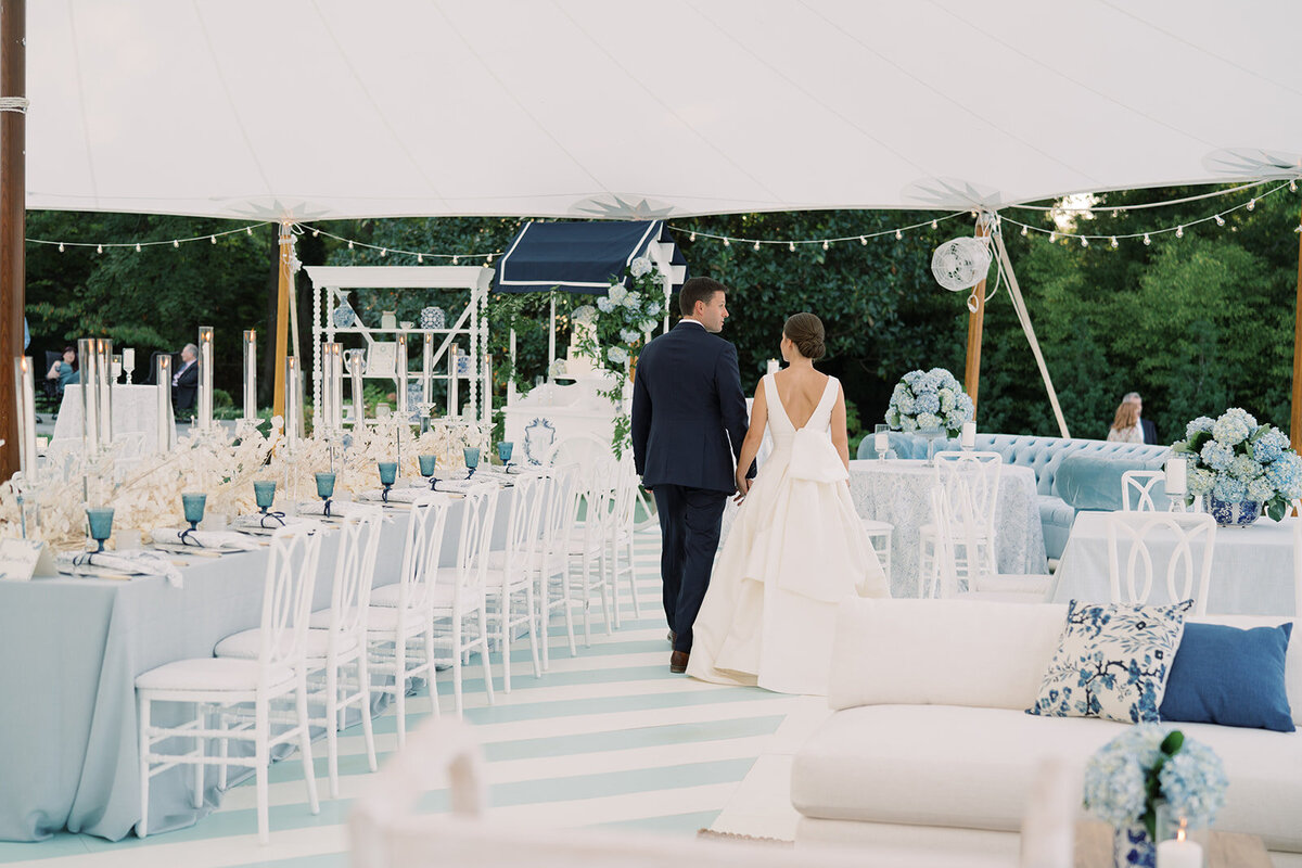 The photo was taken at The Hamptons, New York, waterfront estate tented wedding by photographer Bonnie Sen. In addition, East West Films was the videographers capturing the beautiful moments at the outdoor wedding reception. The venue features white tables, blue glassware, blue and white cushions, and elegant floral arrangements, creating a serene and elegant atmosphere. The waterfront estate offers picturesque views of the ocean, adding a touch of natural beauty to the already charming setting.