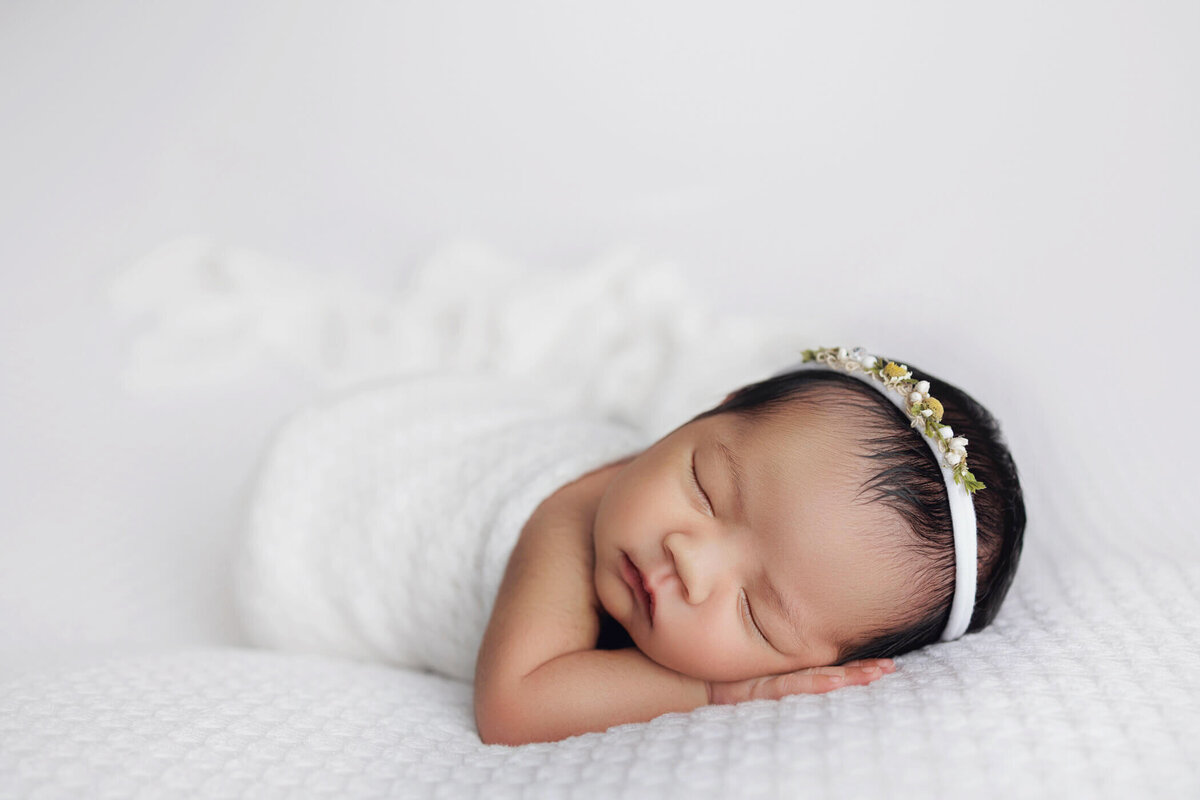 Newborn baby wrapped in a white swaddle, wearing a white headband with a small flower, peacefully sleeping on a white textured blanket.