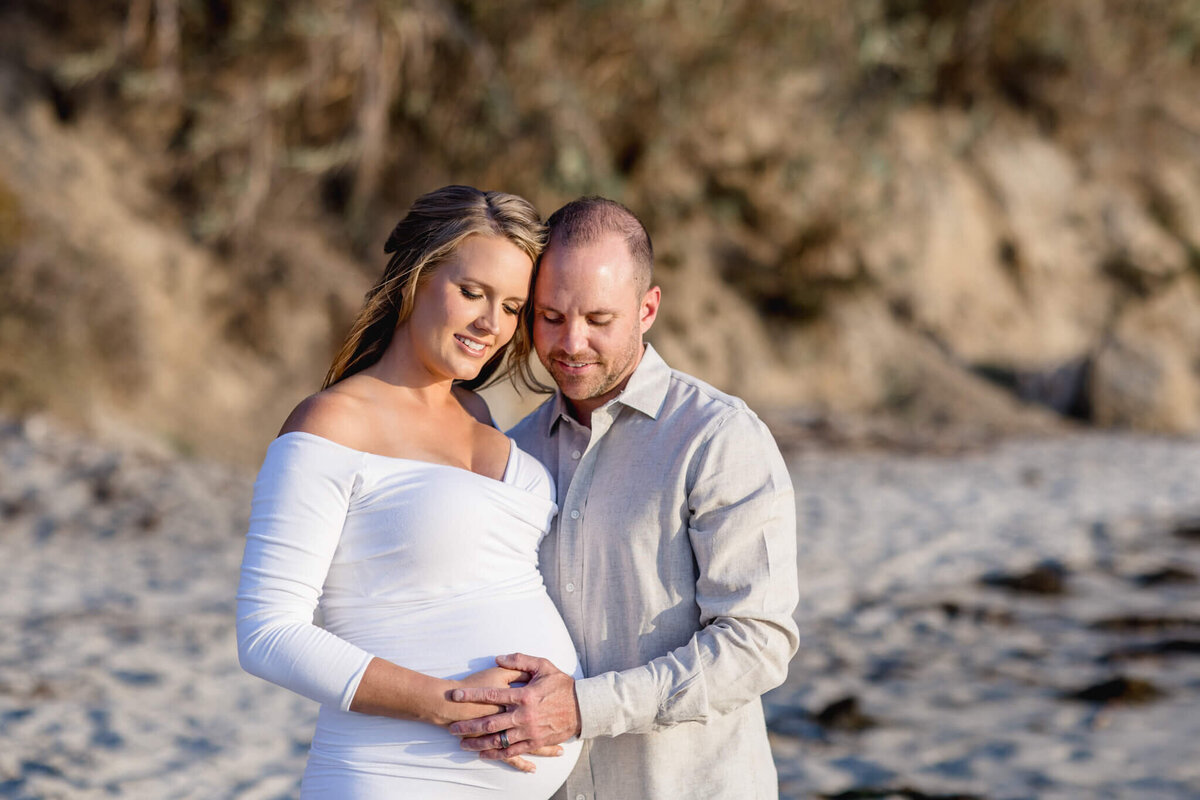 A pregnant woman in a white dress stands on a sandy beach, embraced by a man in a light shirt. They both gently place their hands on her belly, smiling softly. The background features sand and blurred vegetation.