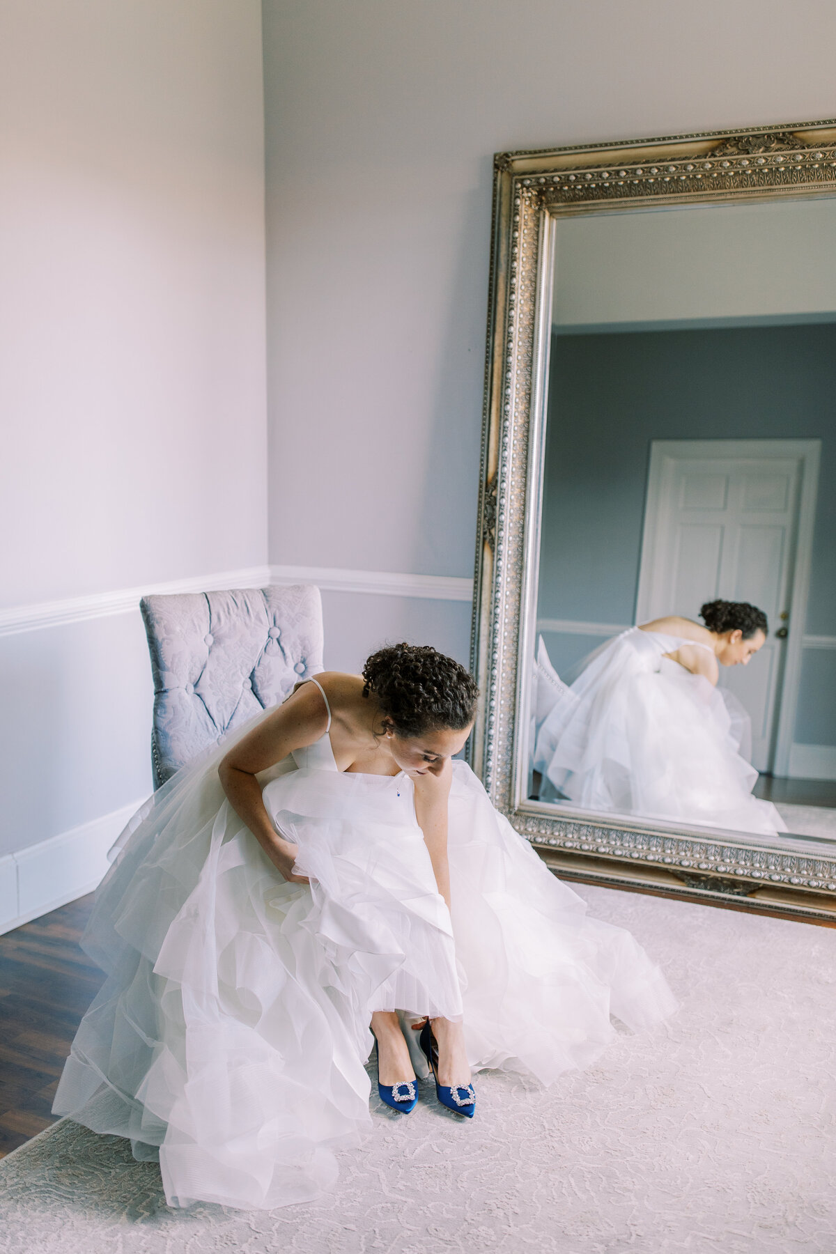 Bride puts on her shoes in a blue room with a long mirror showing her relfection