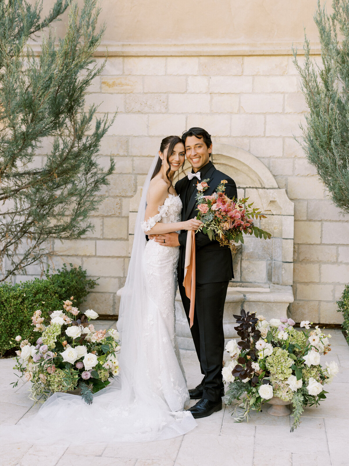 Newlyweds glowing with joy in front of a stone fountain, surrounded by romantic floral arrangements—timeless elegance meets vineyard charm.