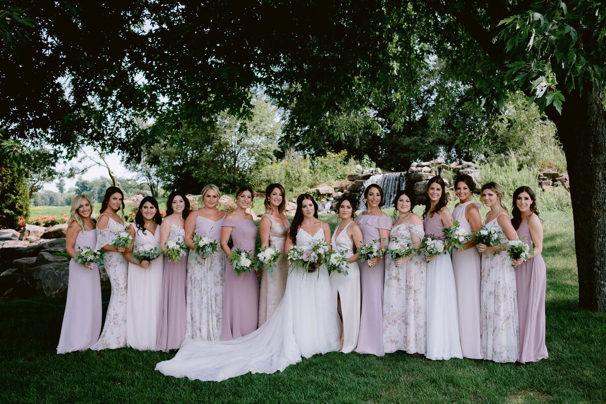 A large bridal party poses in a green Montreal park holding garden flower bouquets.