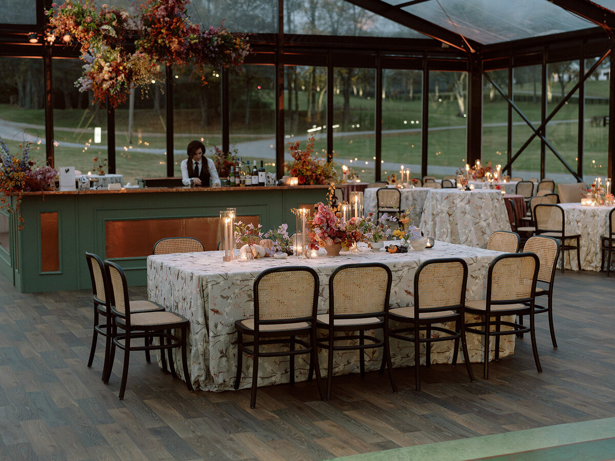 Clustered and styled floral arrangements, fruits, and art pieces for fall wedding reception. Low centerpieces mixed with ikebana arrangements and bud vases decorate the reception tables at this fall wedding. Floral centerpieces featuring orchids, ranunculus, sweet peas, and bittersweet. Styled wedding tables with fruits, curated candles, and floral arrangements. Tasteful fall color palette of copper, ruby, gold, burgundy, rust, and lavender. Design by Rosemary & Finch Floral Design in Nashville, TN.