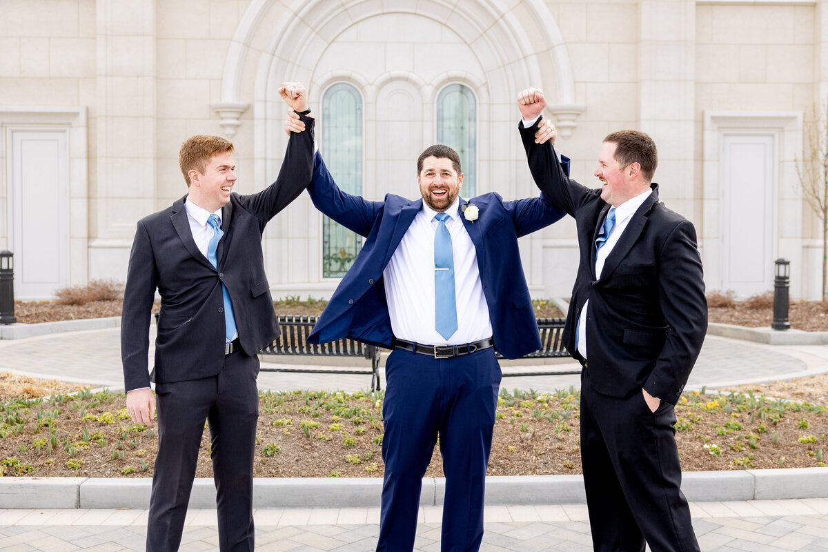Groom holds his arms up joyfully as his two brothers lift his hands in celebration, all smiling in front of the temple.