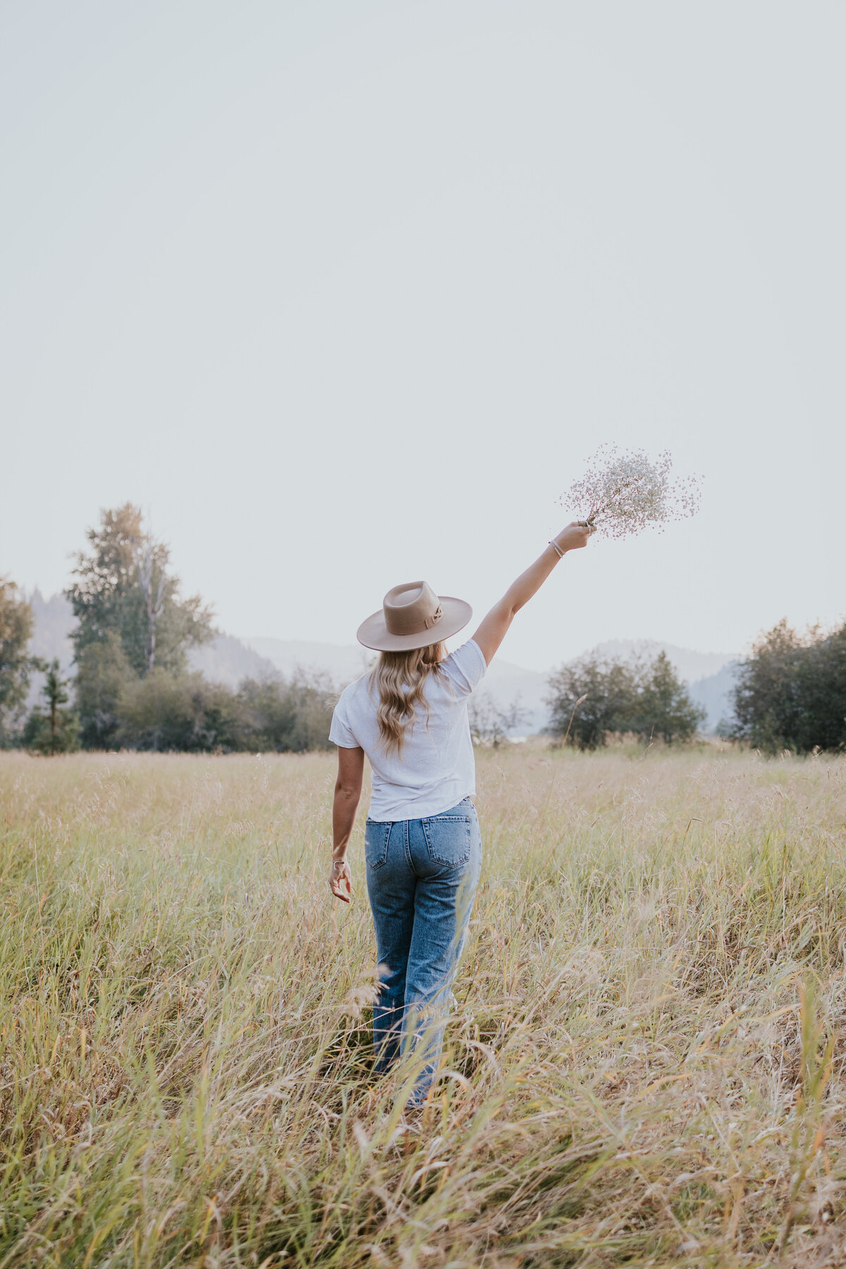 Young woman walks away from camera in field while swinging bouquet of flowers above her head.