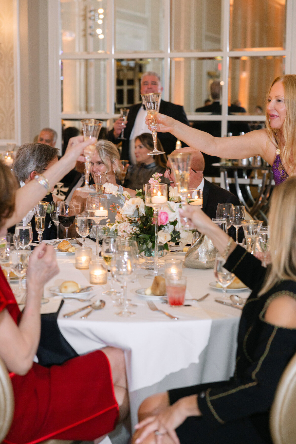 guests toasting with glasses with champagne at wedding reception elegantly styled wedding reception table at The Adolphus in Dallas featuring a black tablecloth, tall glass vases with pink rose arrangements, numerous candles, and refined glassware and chargers 