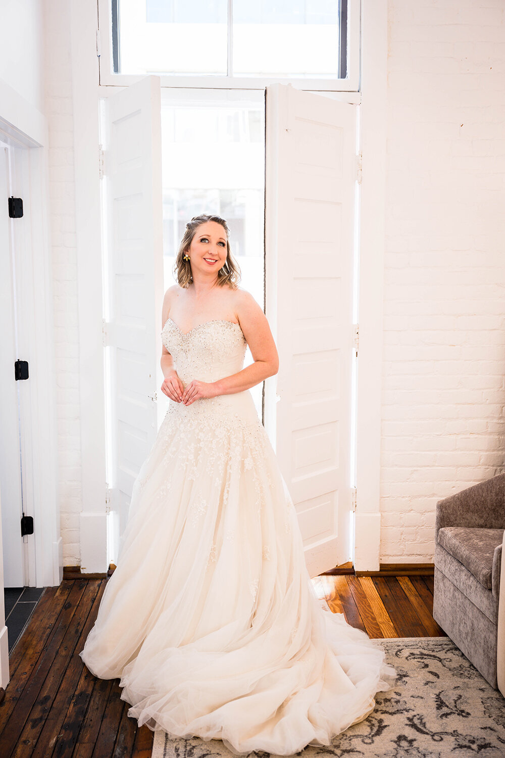 A bride stands in front of a window with large doors inside a Fire Station One Boutique Hotel room in Downtown Roanoke, Virginia for a formal portrait.
