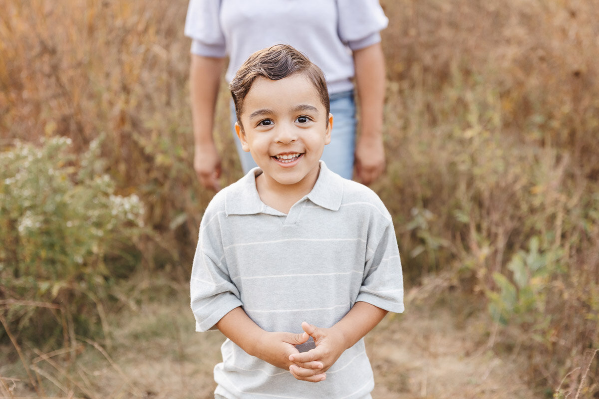 (direct upload) A young child with short hair smiles at the camera, with an adult partially visible in the background, capturing a tender moment. This candid shot by a family photographer in Toronto perfectly highlights the charm of a dry, grassy setting.