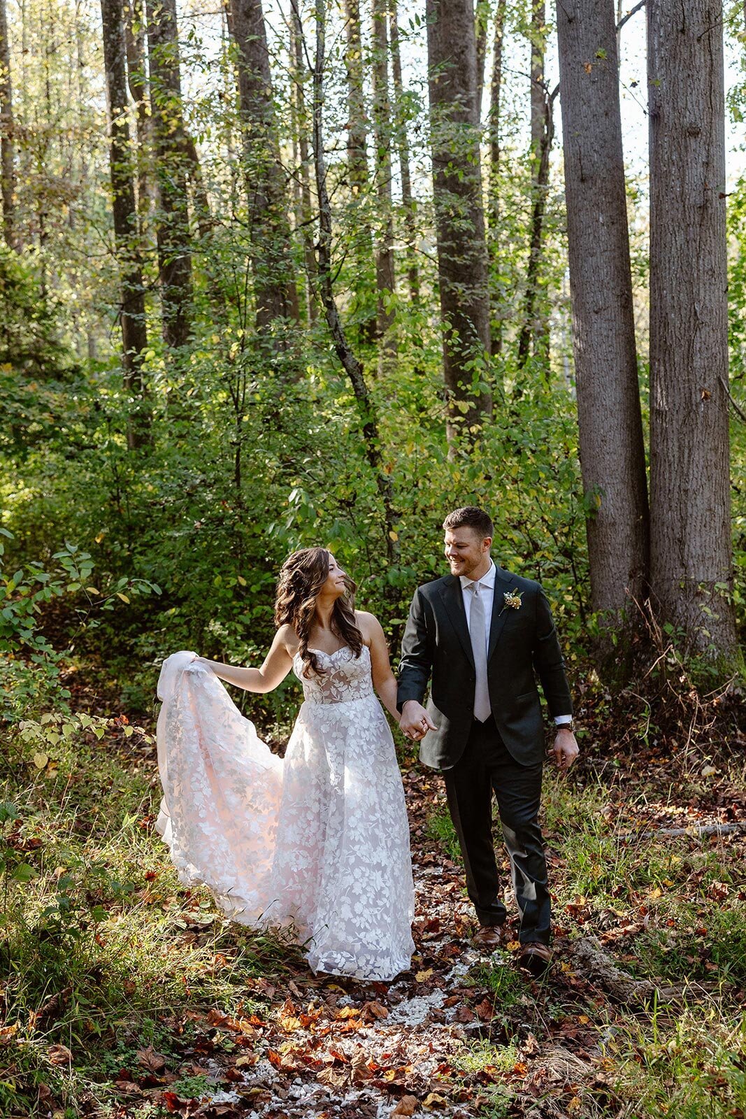 A bride and groom walking in the woods on their wedding day. They are looking at one another and smiling.