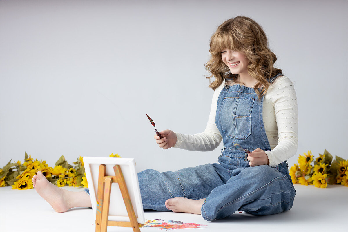A young woman wearing denim overalls and a white long-sleeve shirt smiles while holding a paintbrush. She sits on the floor surrounded by sunflowers and colorful paint splashes, focused on her small canvas. Her relaxed posture and joy reflect her love for painting in this creative moment.