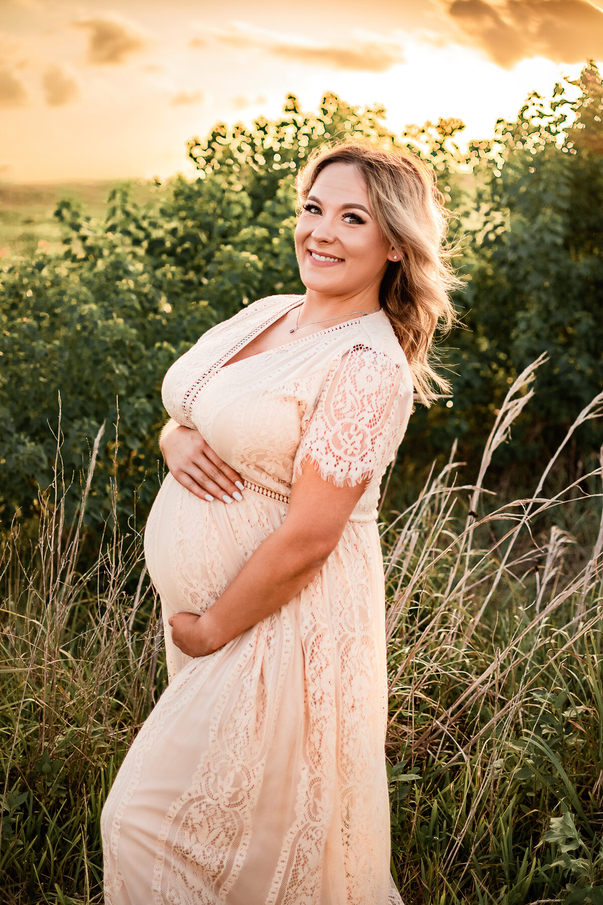 A woman wearing a tan dress holds the top and bottom of her baby bump and smiles at the camera.